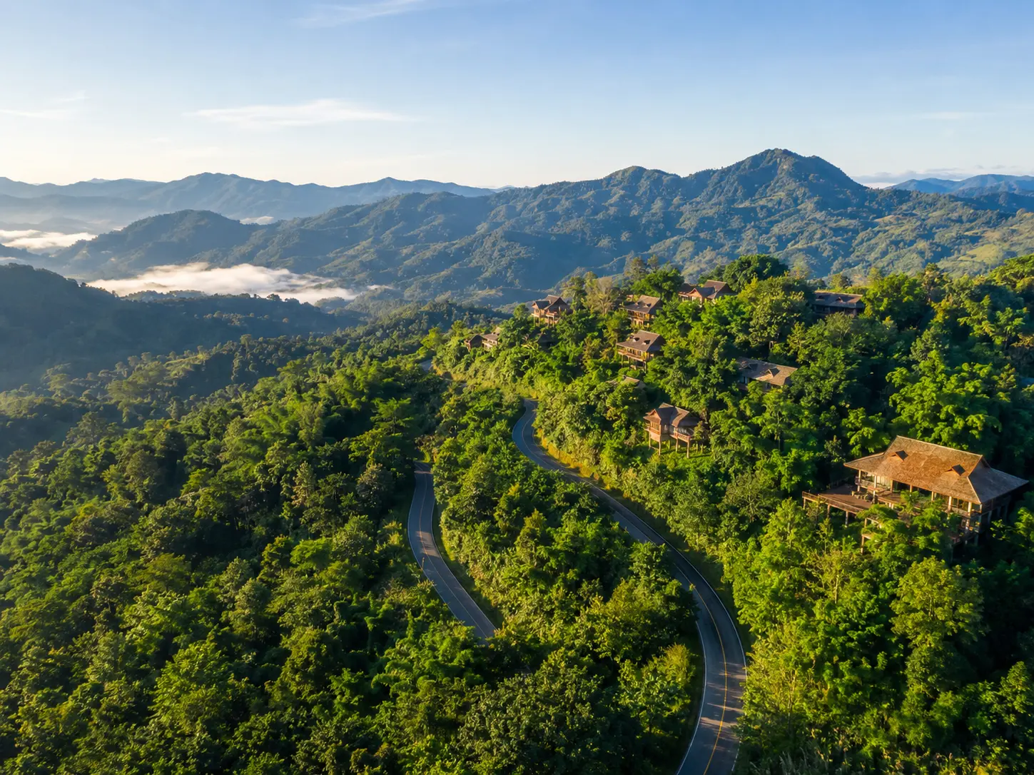 Bright aerial view of mountain cabins and a winding road surrounded by lush green forest in northern Thailand